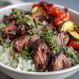 Freshly grilled steak slices rest atop fluffy white rice with colorful roasted vegetables and chimichurri in a warm bowl.